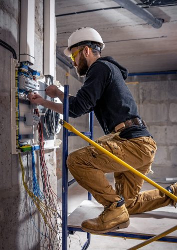 A male electrician works in a switchboard with an electrical connecting cable, connects the equipment with tools.